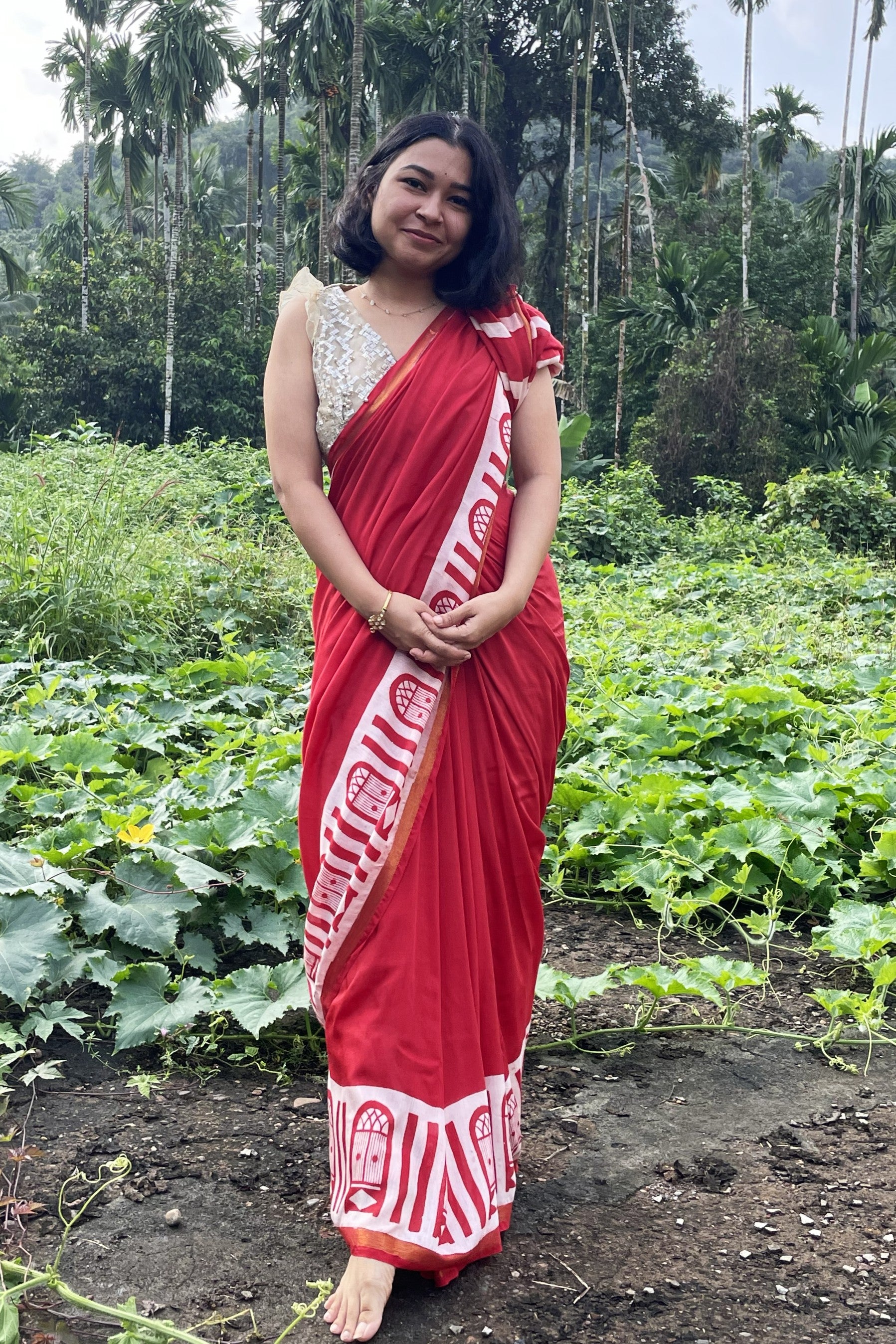 Woman in a red Mogasu saree standing in a lush green field with trees in the background