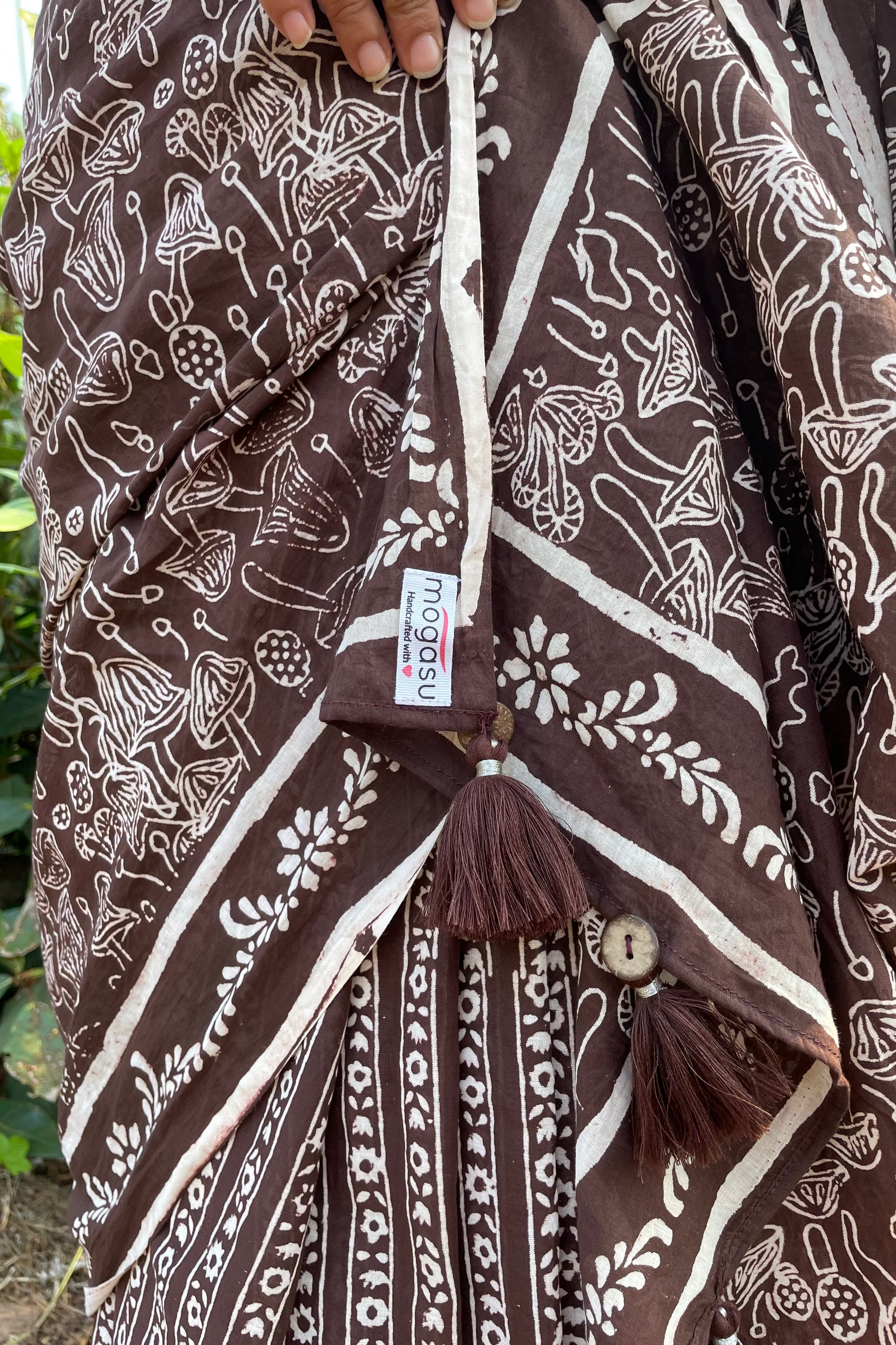 Brown and white mushroom patterned saree with a Mogasu brand label, held by a hand against a natural background in Goa.