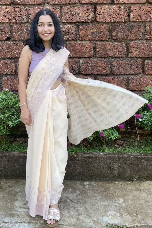 Woman in a Mogasu cream mul cotton saree standing against a brick wall with greenery.