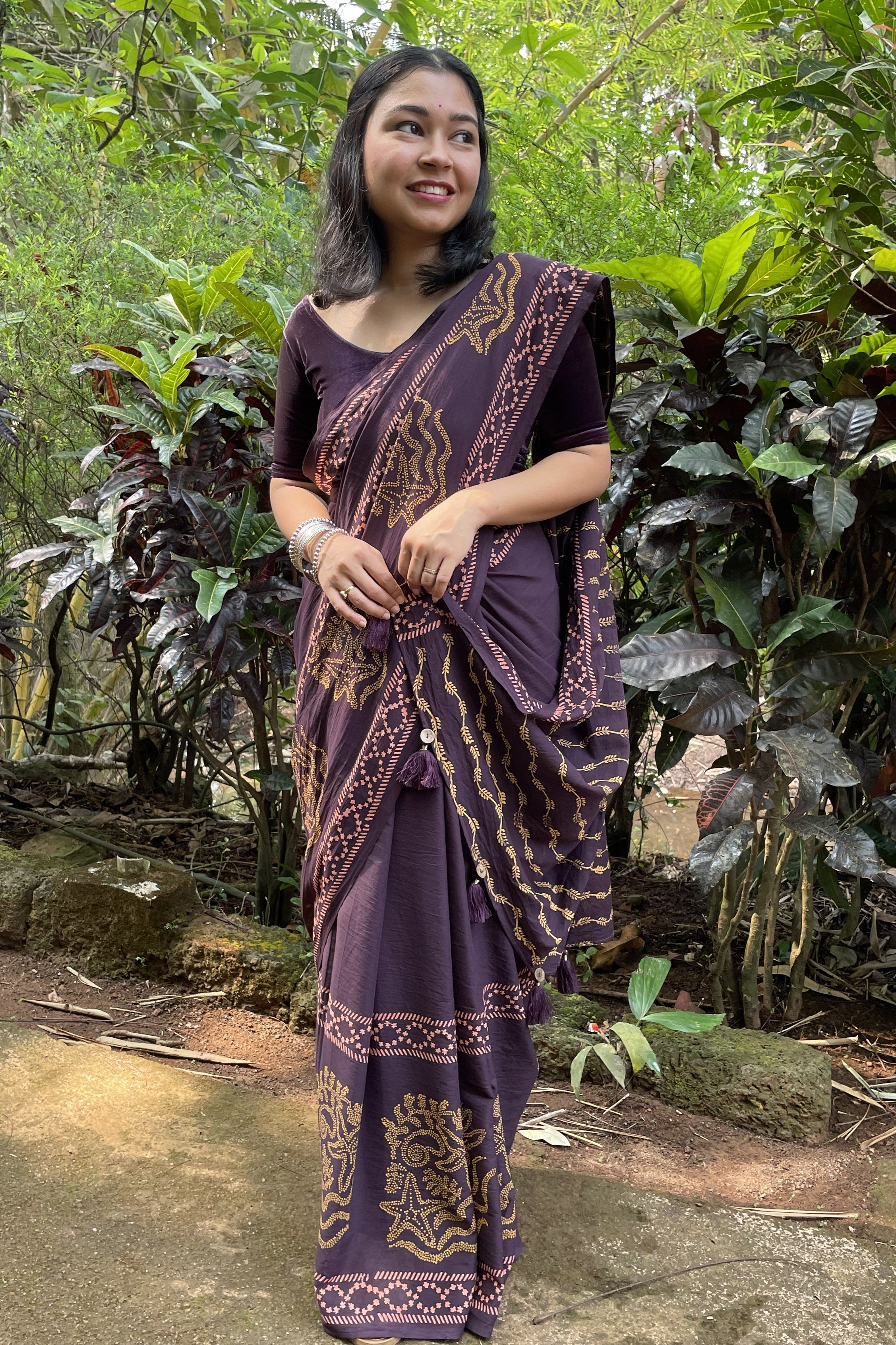 Woman in a traditional purple and gold saree standing in a forest setting in Goa. She as draped Mogasu’s mul cotton saree.