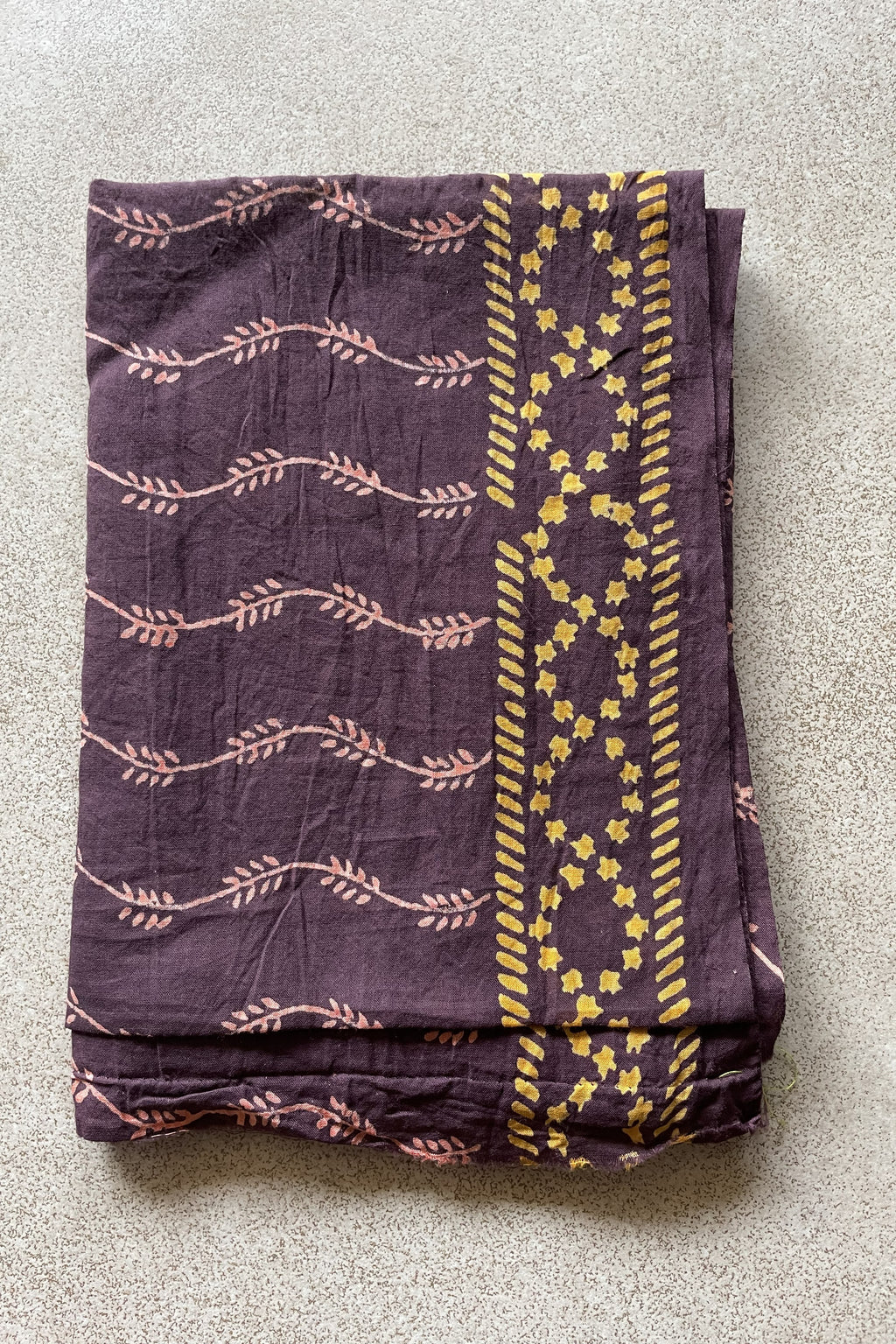 Folded purple blouse piece with yellow patterns on a gray surface. Mogasu’s blouse piece for Noa saree in mul cotton.