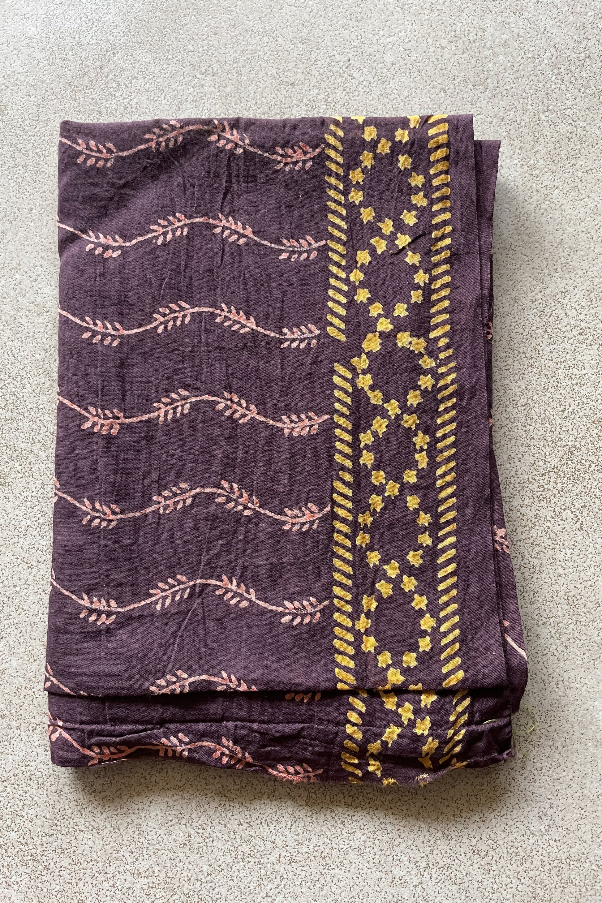 Folded purple blouse piece with yellow patterns on a gray surface. Mogasu’s blouse piece for Noa saree in mul cotton.