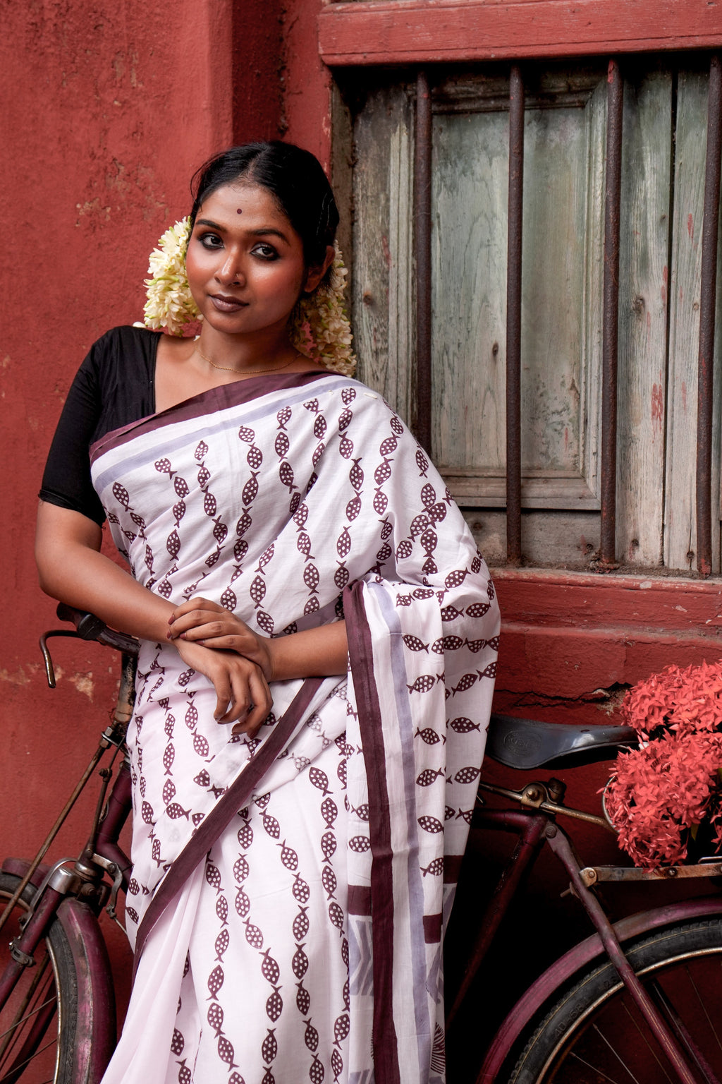 Woman in a light pink Mogasu saree with brown patterns standing next to a bicycle against a red wall.