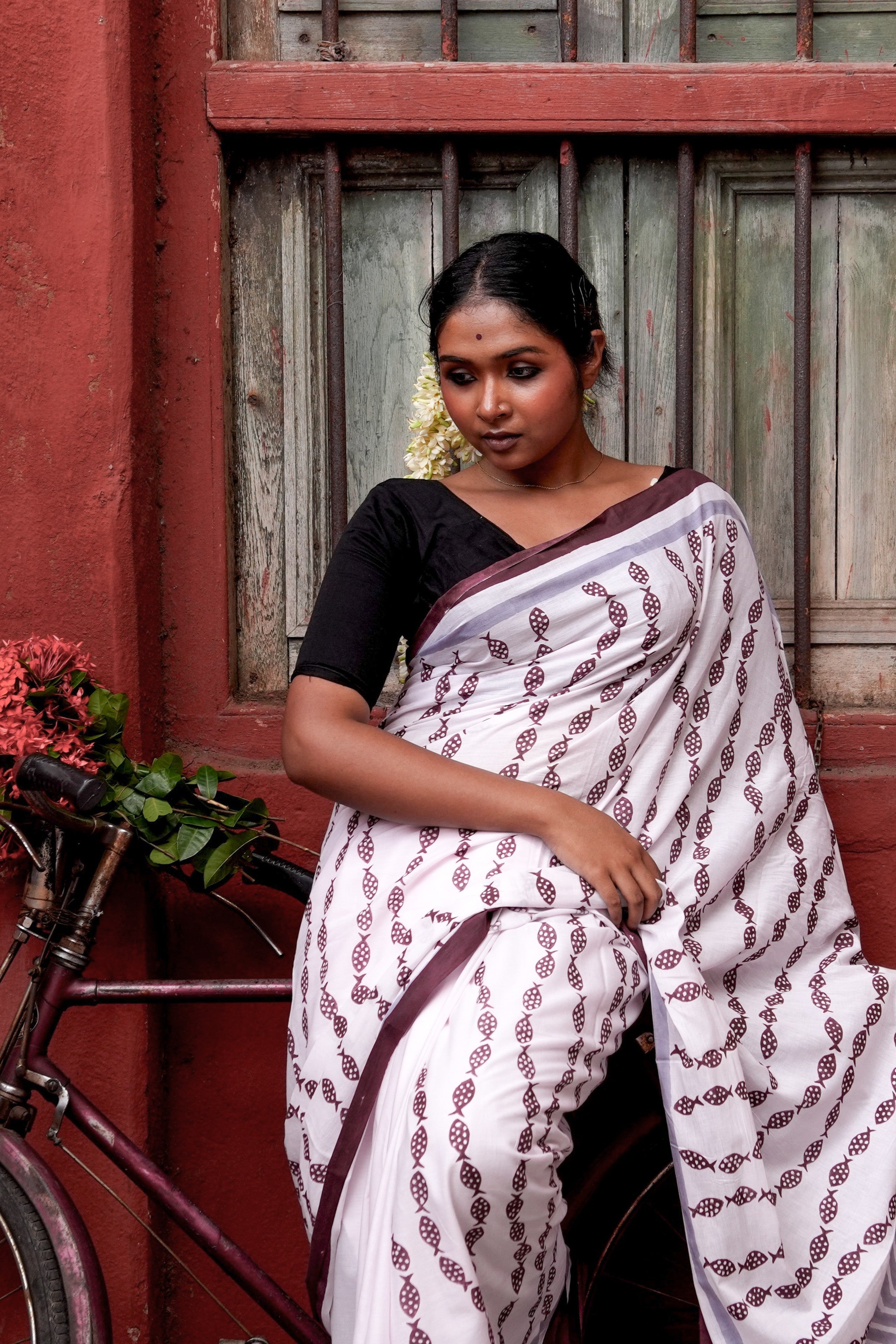 Woman in a light pink Mogasu saree with red patterns standing next to a bicycle against a red wall.