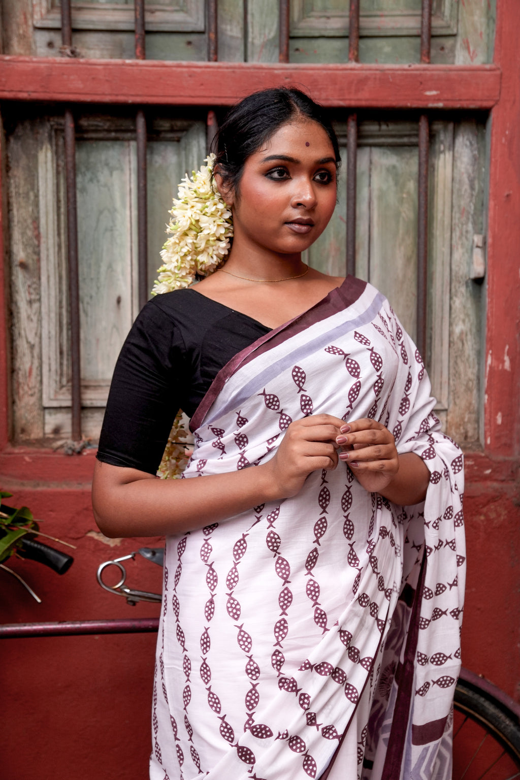 Woman wearing a light pink Mogasu saree with a red border in front of a wooden door.