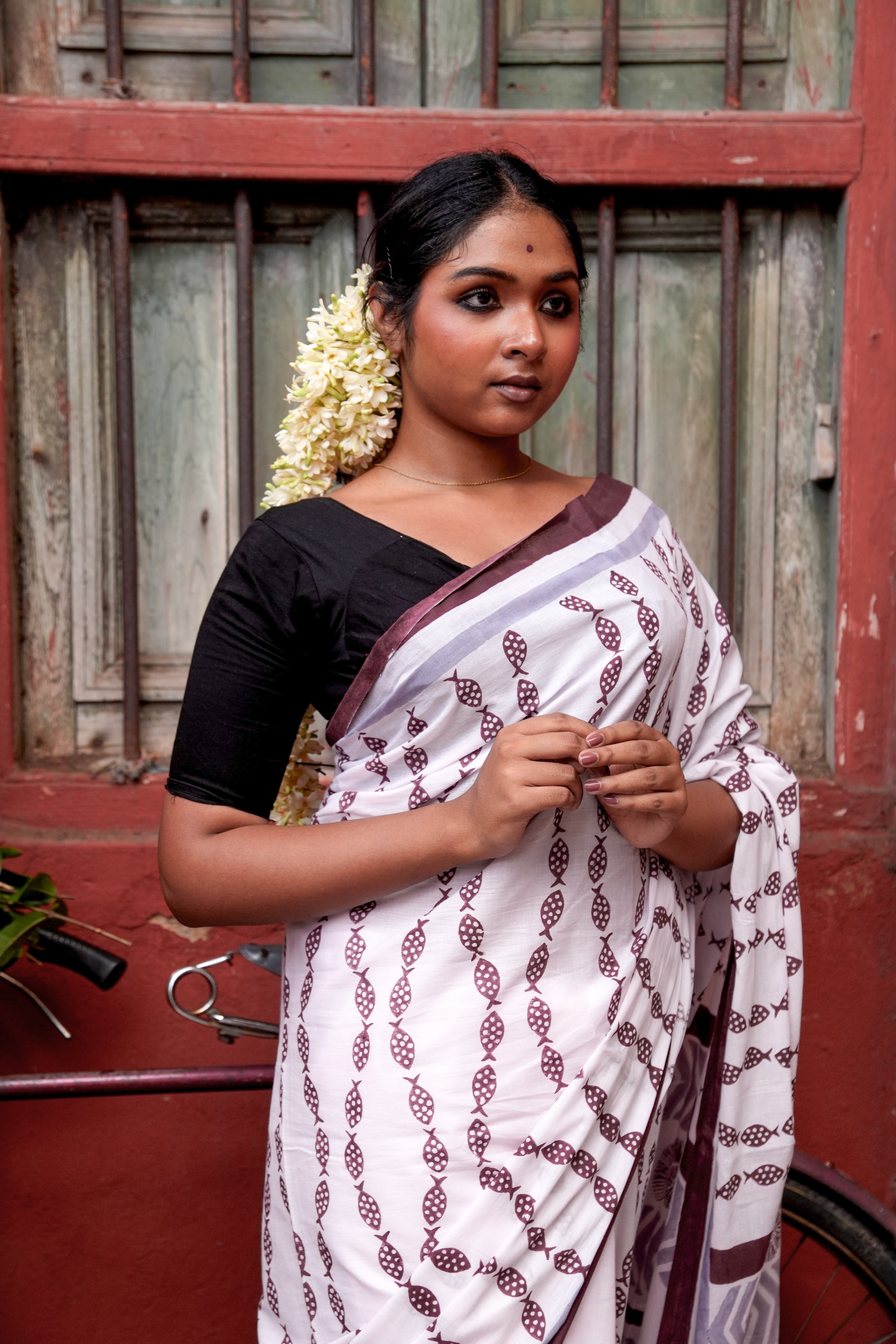 Woman wearing a light pink Mogasu saree with a red border in front of a wooden door.