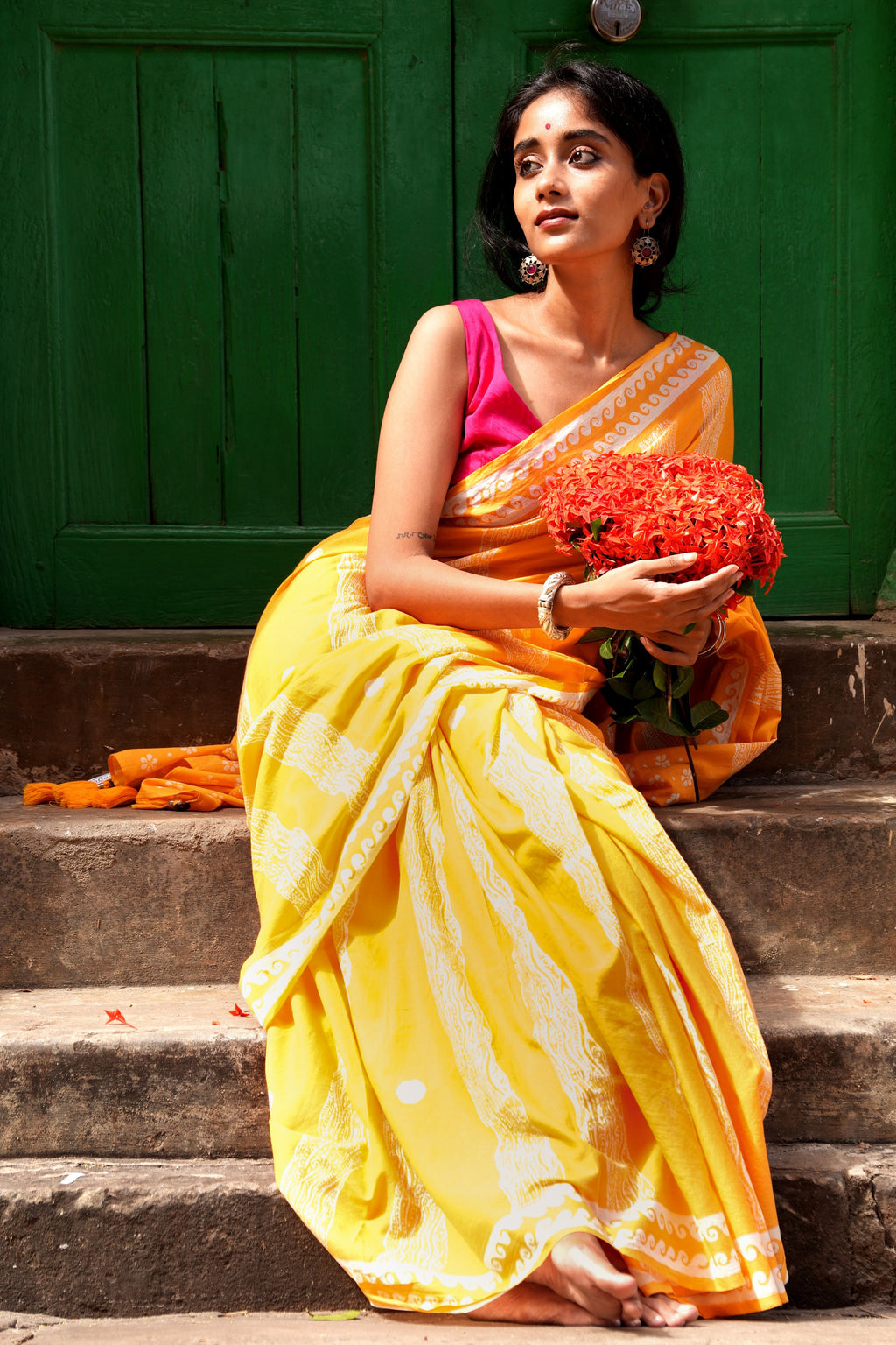 Woman in a yellow Mogasu Brisa saree with a pink blouse holding flowers, sitting on steps against a green door.