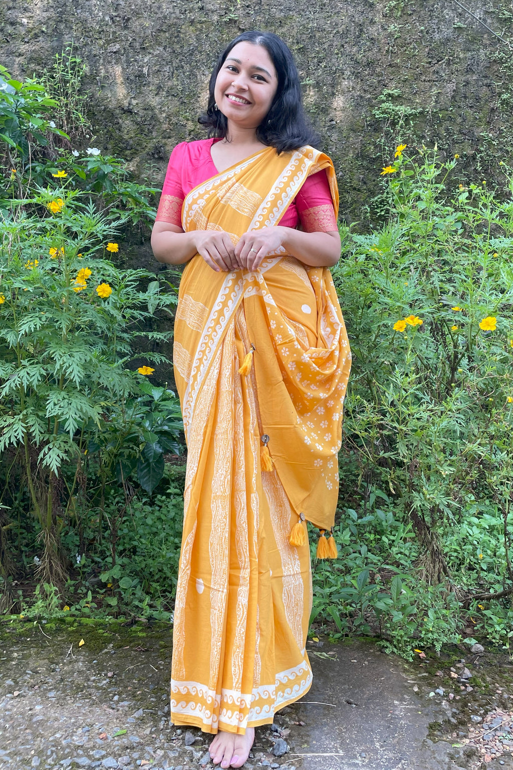 Woman in a yellow Mogasu mul cotton saree with a pink blouse standing outdoors among greenery.