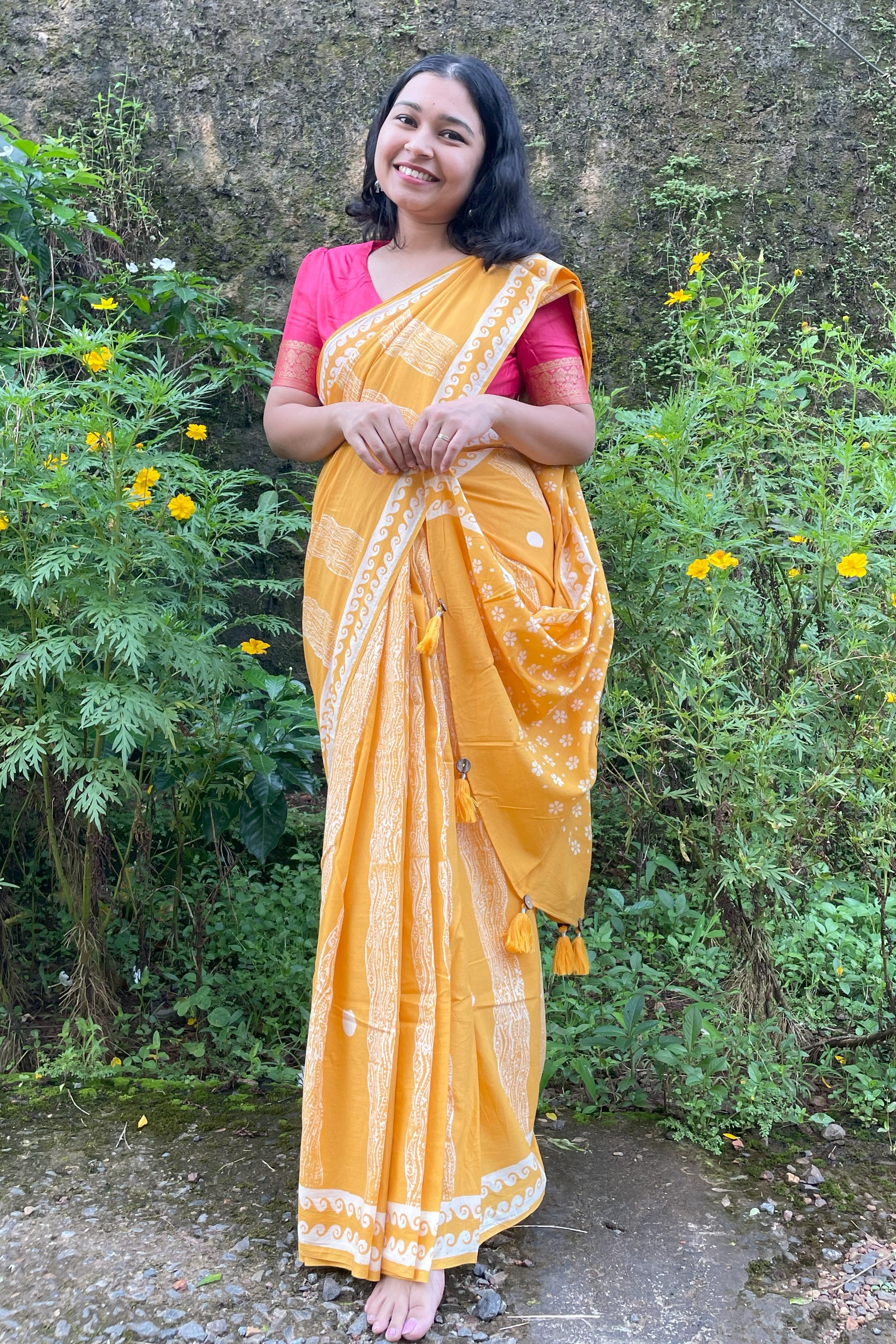 Woman in a yellow Mogasu mul cotton saree with a pink blouse standing outdoors among greenery.