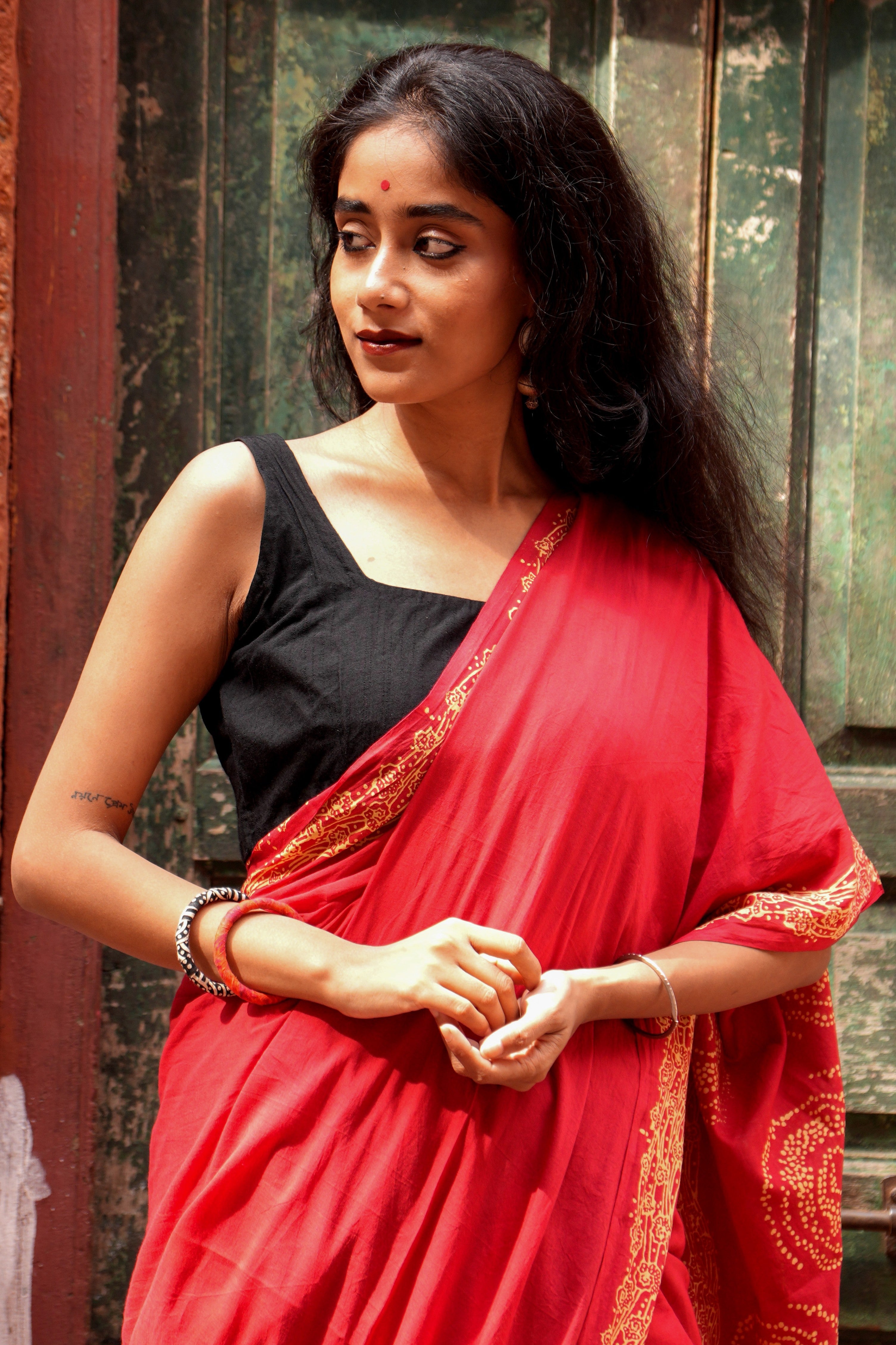 Woman wearing a red Mogasu mul cotton saree with a black blouse against a textured wall.