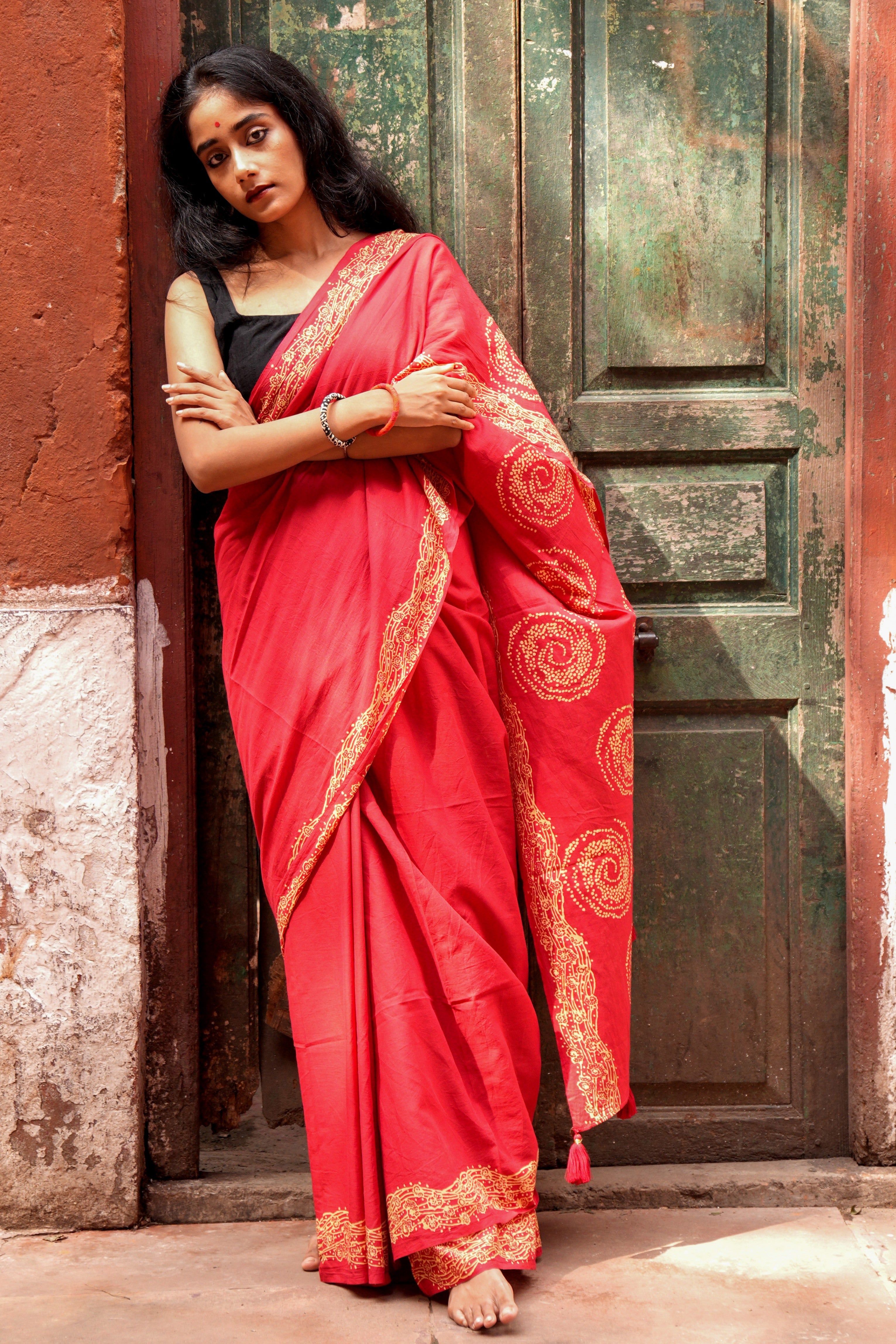 Woman in a red Mogasu Lhaar saree with gold patterns standing against a textured wall.