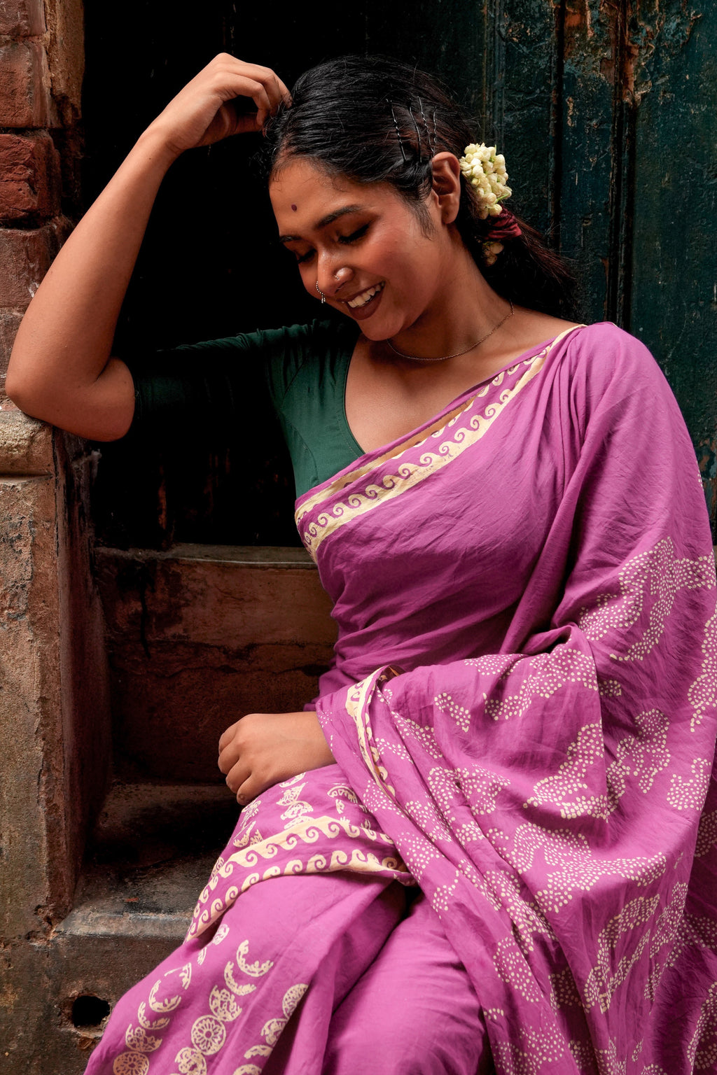 Woman in a purple Mogasu saree with green blouse sitting against a rustic wall.