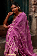 Woman wearing a purple Mogasu Lua saree with gold patterns, standing against a dark wall.