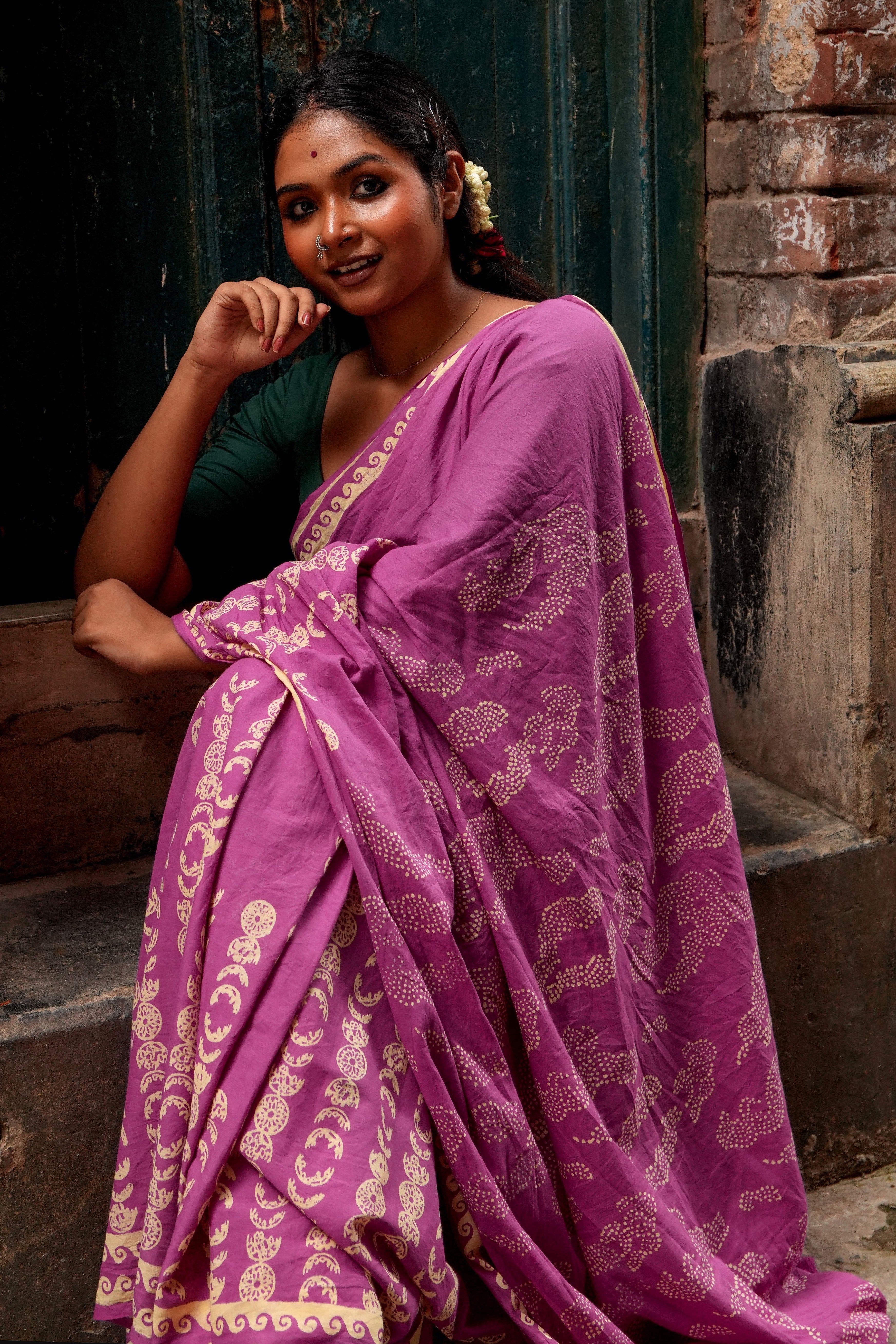 Woman wearing a purple Mogasu Lua saree with gold patterns, standing against a dark wall.