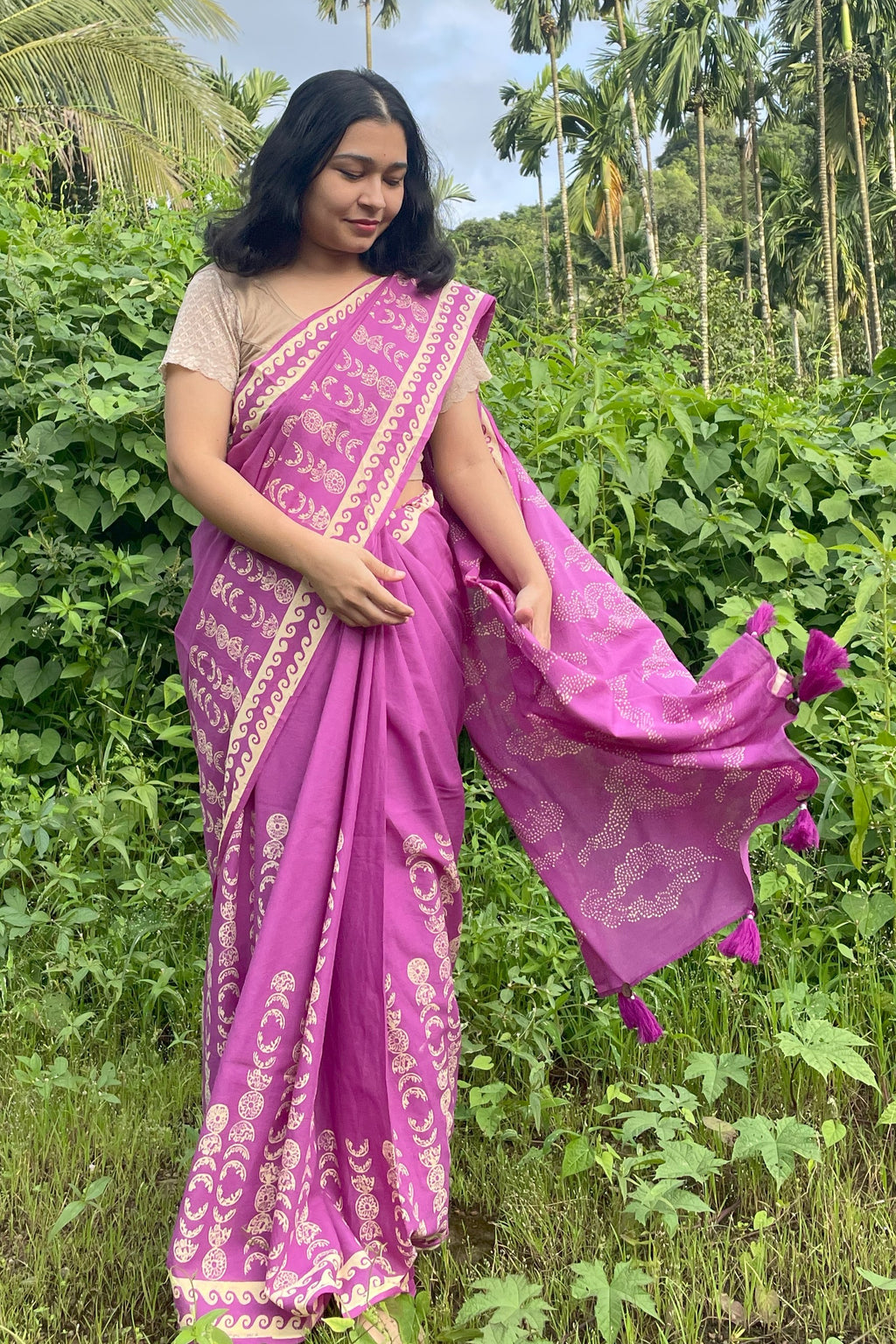 Woman in a purple Mogasu saree with gold patterns standing in a lush green outdoor setting in Goa