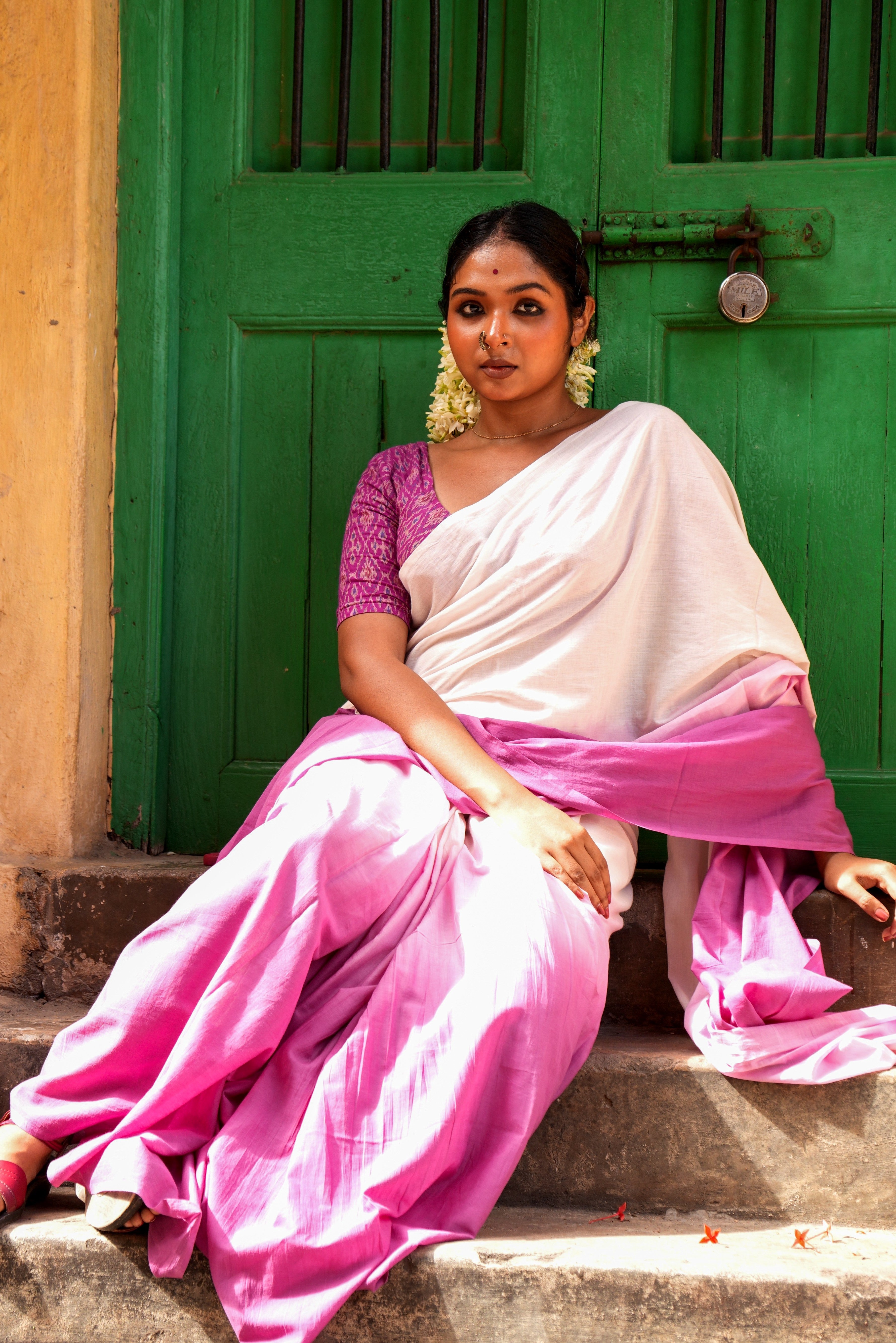 Woman in a pink and white Mogasu saree sitting in front of a green door.