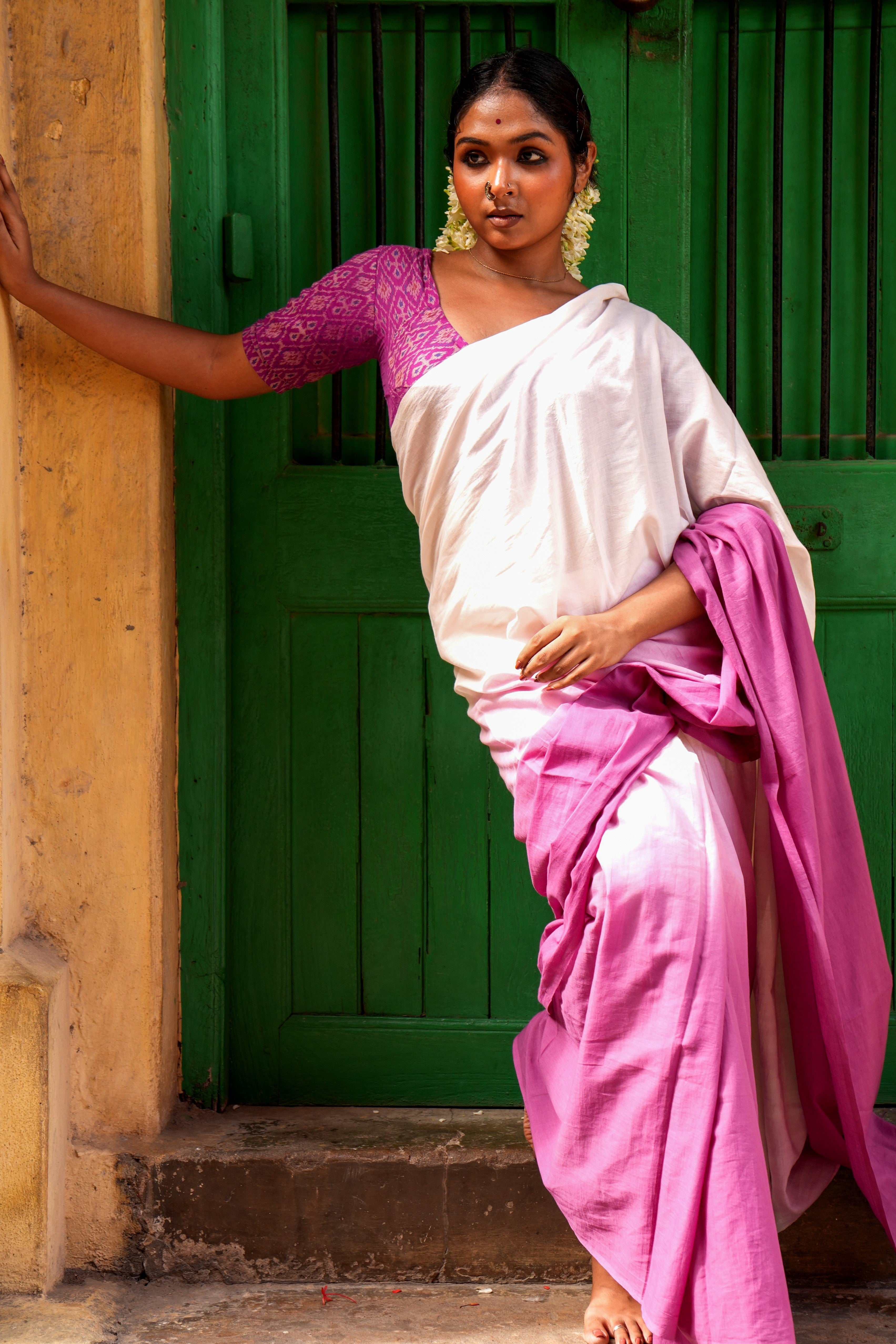 Woman in a white and pink Mogasu Ora mul cotton saree standing against a green door.