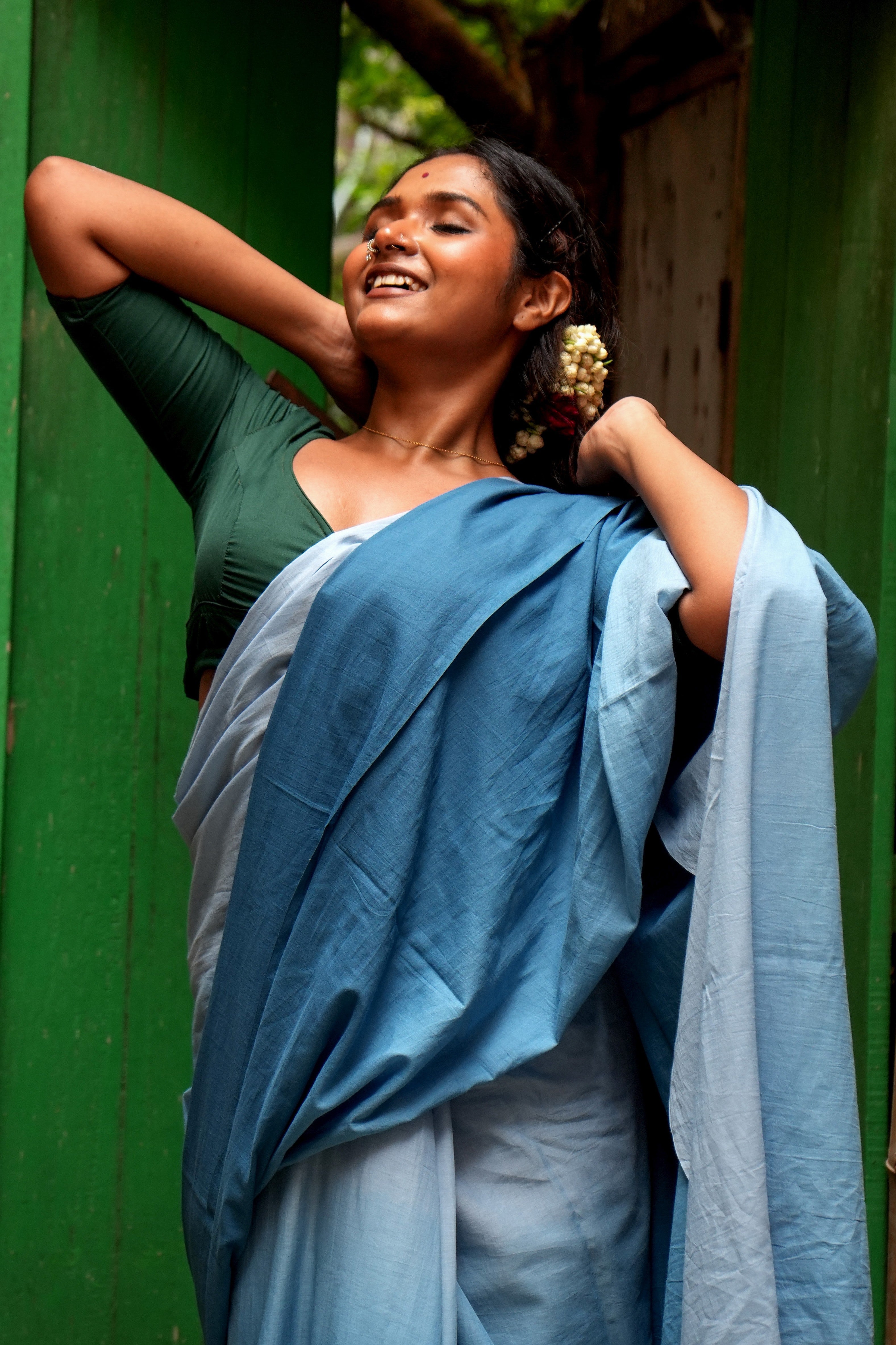 Woman in a Mogasu Rati blue saree standing against a green wall