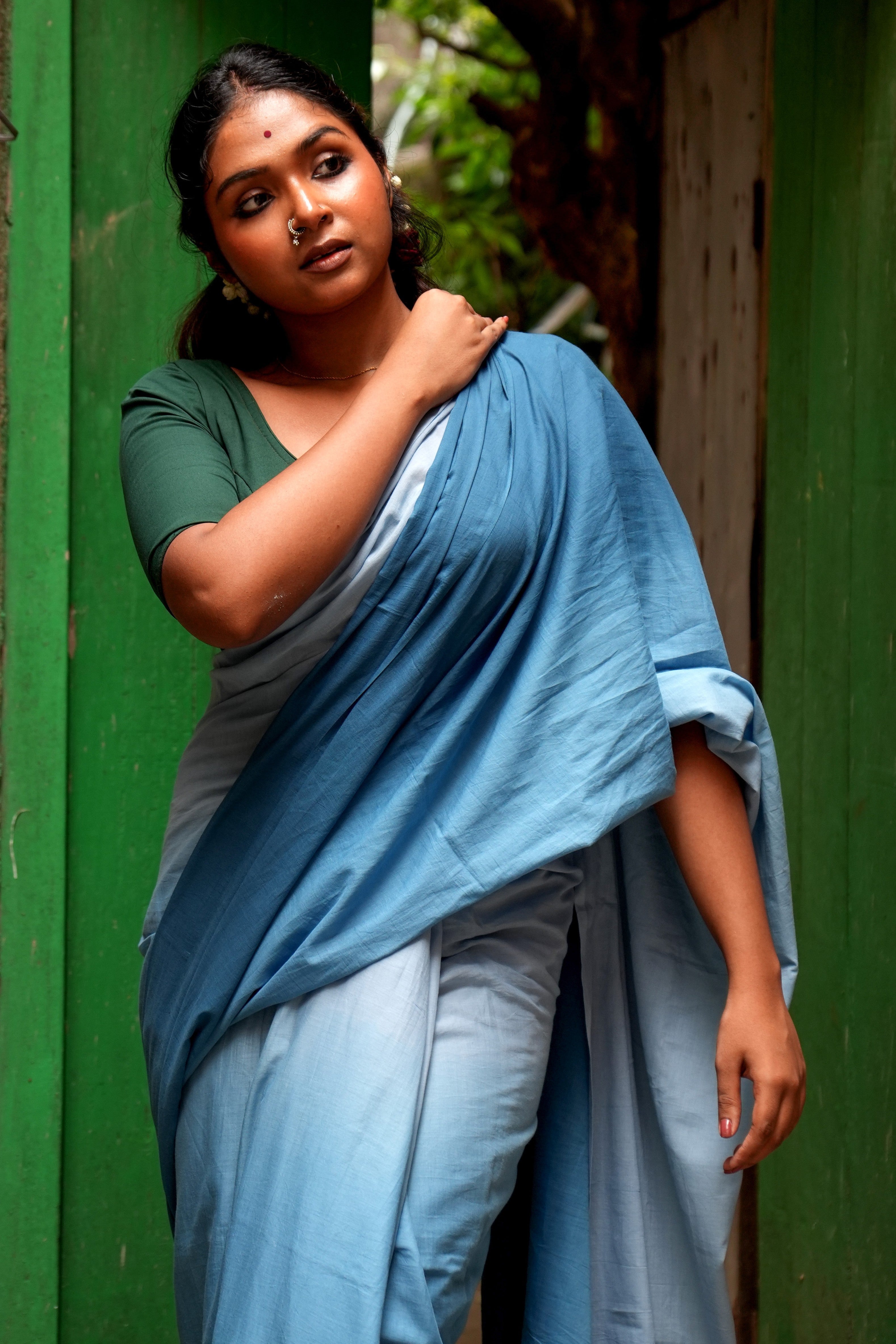 Woman in a Mogasu Rati blue saree standing against a green wall