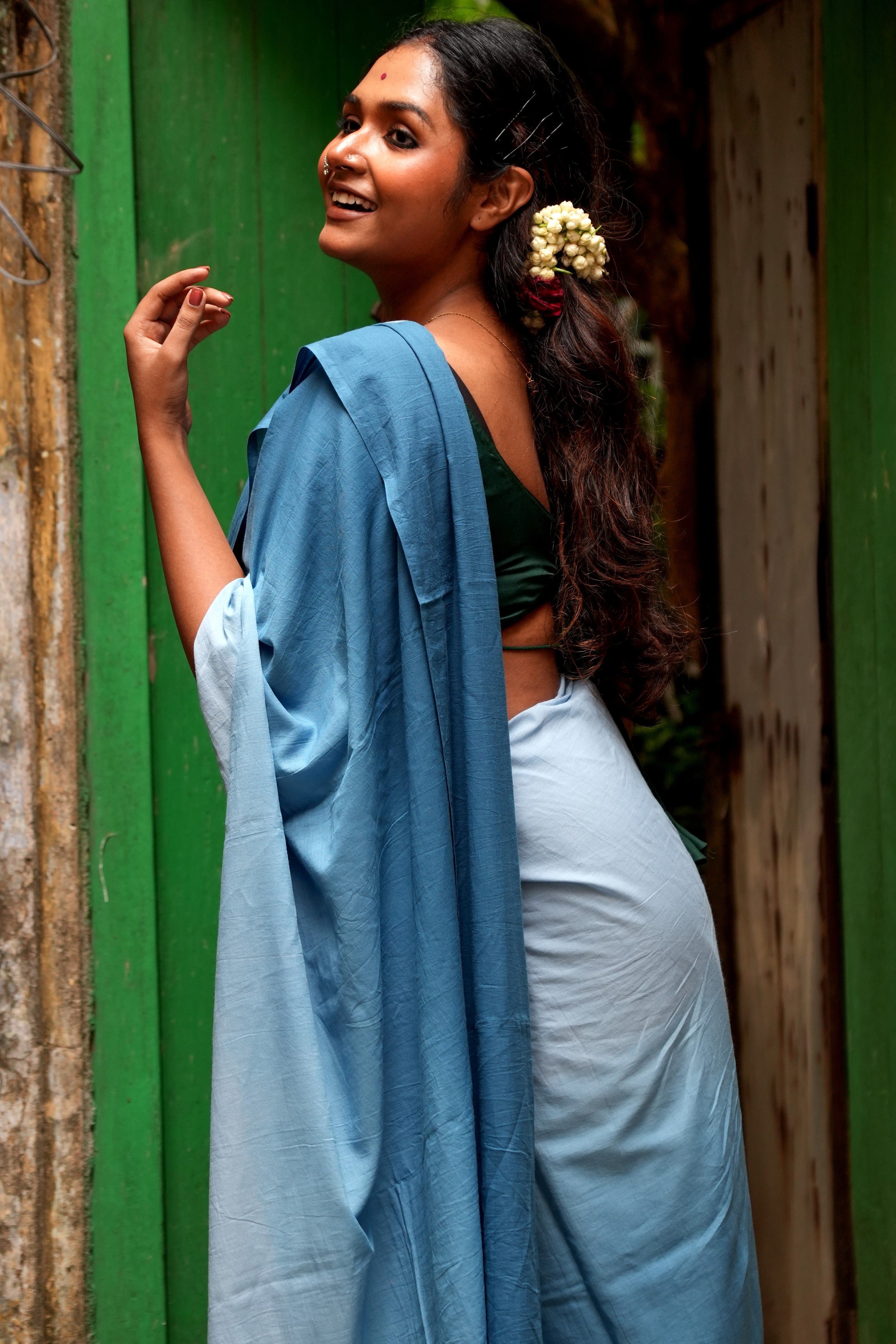 Woman in a Mogasu Rati blue saree standing against a green wall