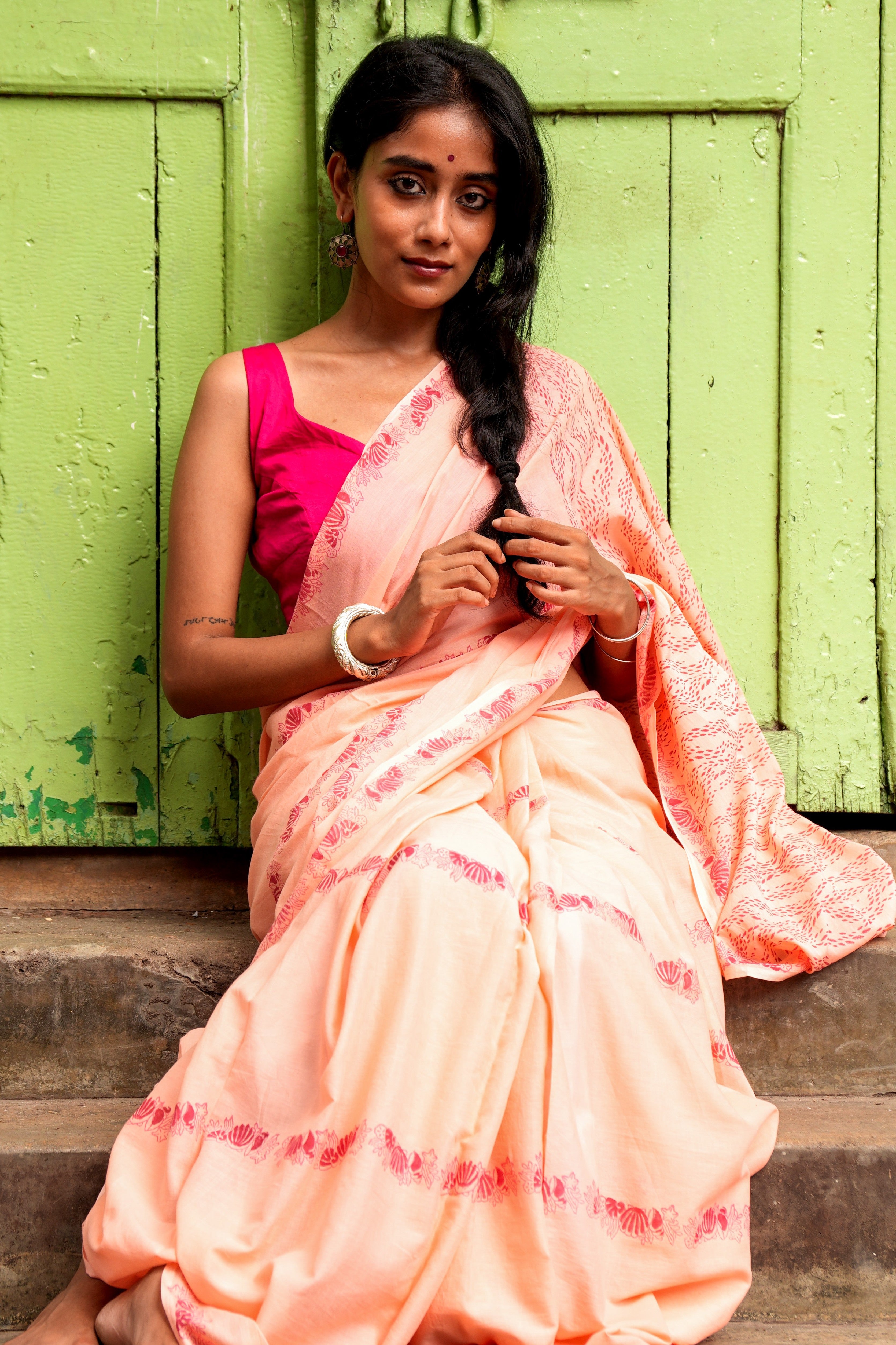 Woman in a peach Mogasu mul cotton saree with pink blouse sitting on steps against a green wooden door.