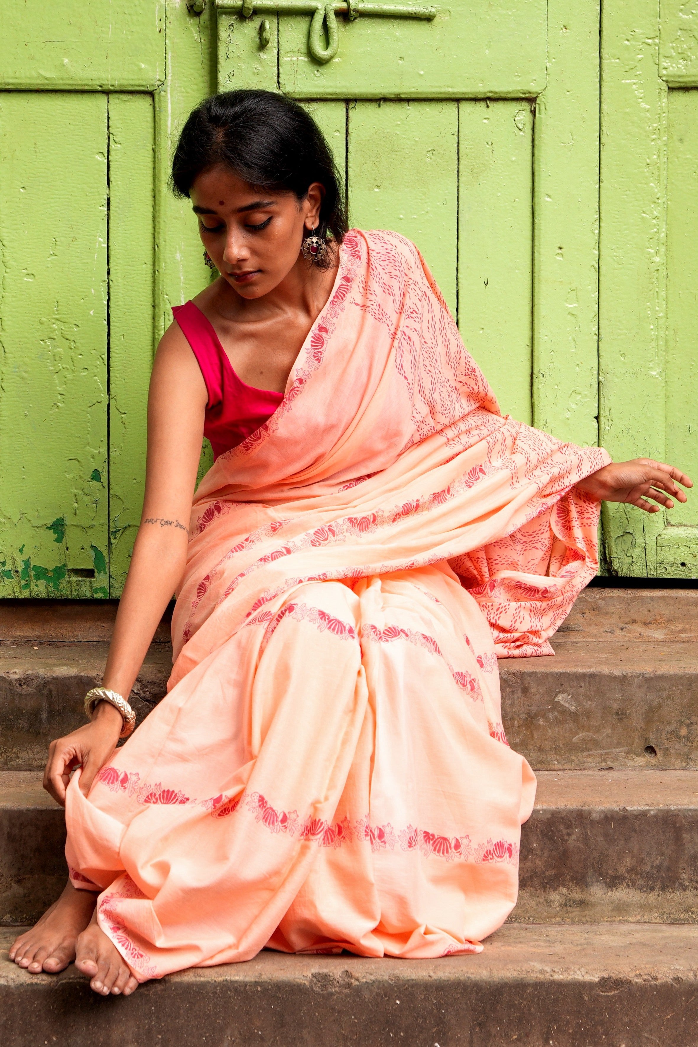 Woman in a peach Mogasu mul cotton saree sitting on steps with a green door in the background