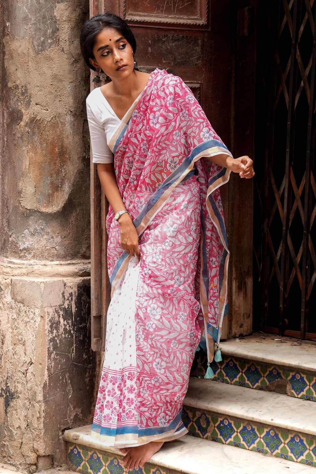 Woman in a pink and white Mogasu saree standing on steps with a textured wall and door in the background