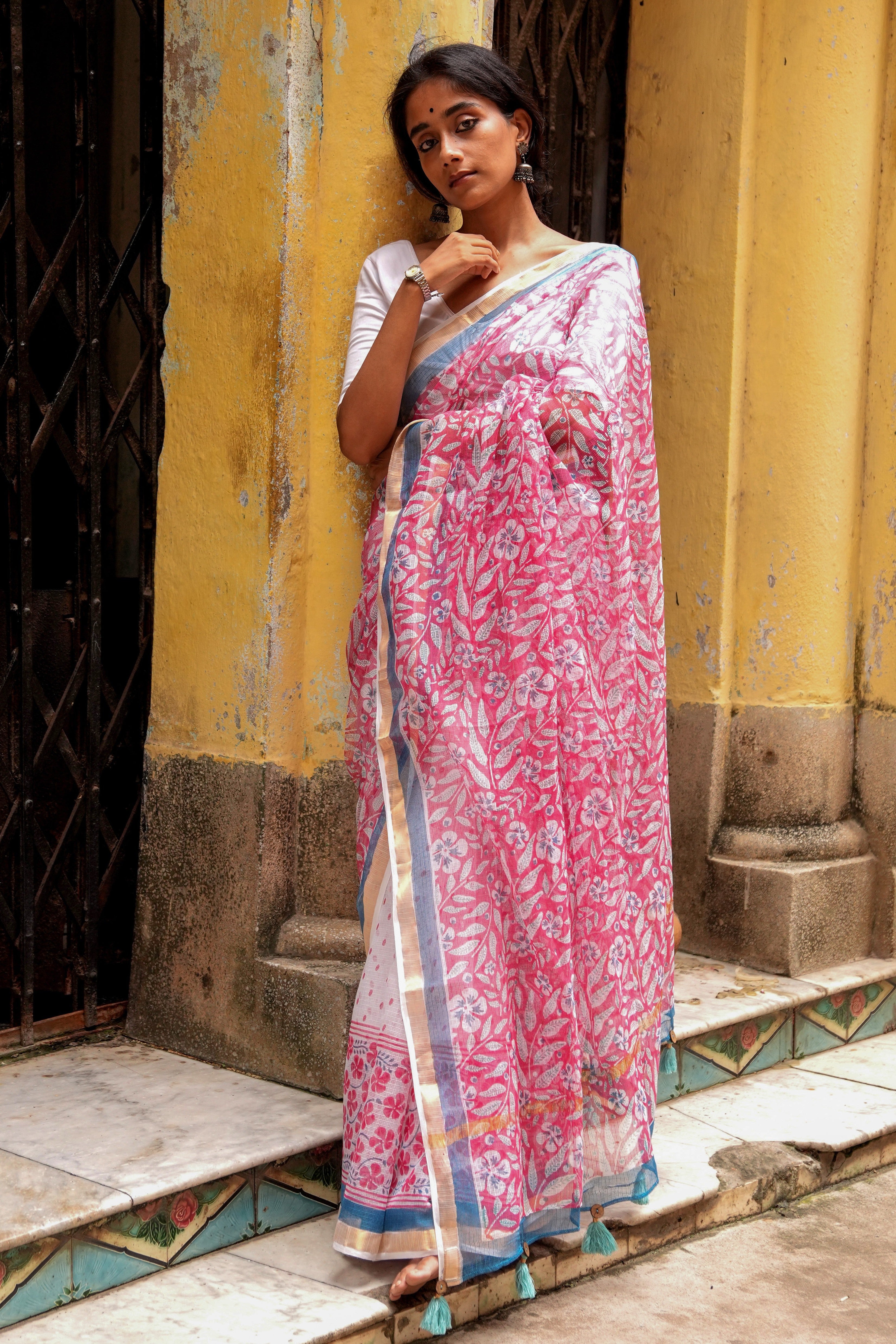 Woman wearing a pink Mogasu kota doria saree with a white background leaning against a yellow wall.
